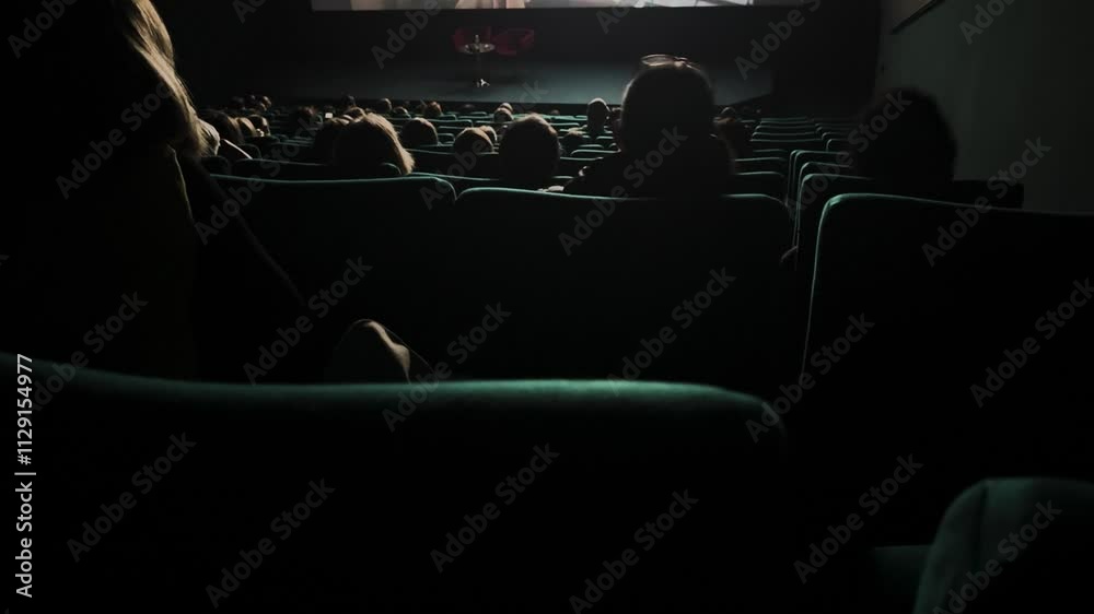 Audience dimly lit movie theater. Empty stage, anticipation. Plush teal seats. Film events premiere. People sit cinema watching at screen. Many spectators dark hall. Back rear view. Evening leisure.