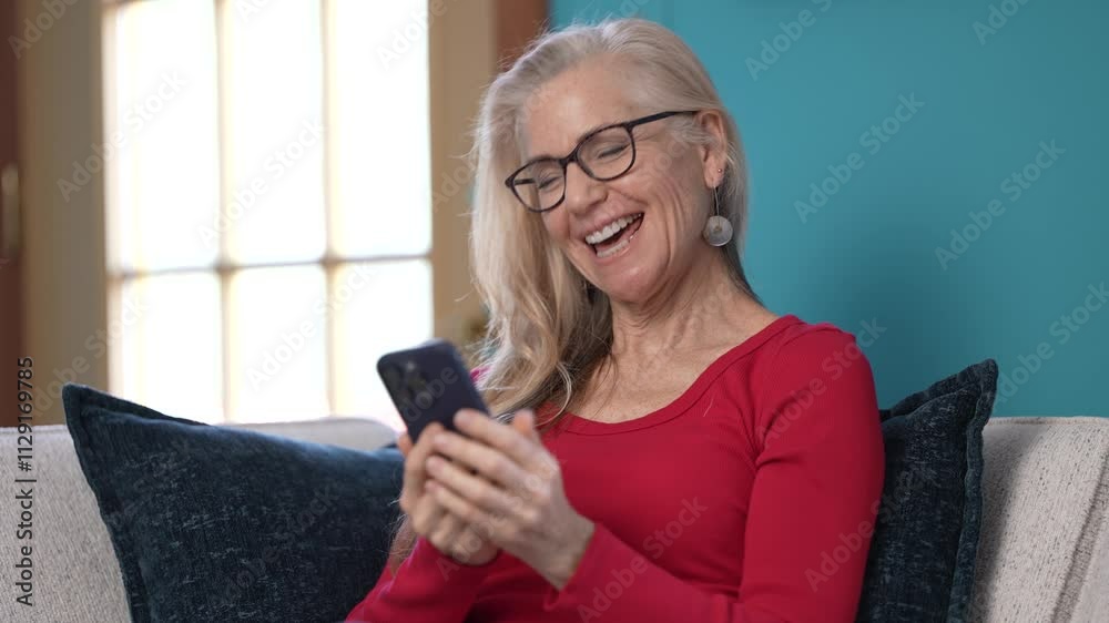 A middle-aged woman with long silver hair enjoys a video call while sitting on a comfortable couch. She wears a red top and displays a joyful expression as she waves to someone on screen.