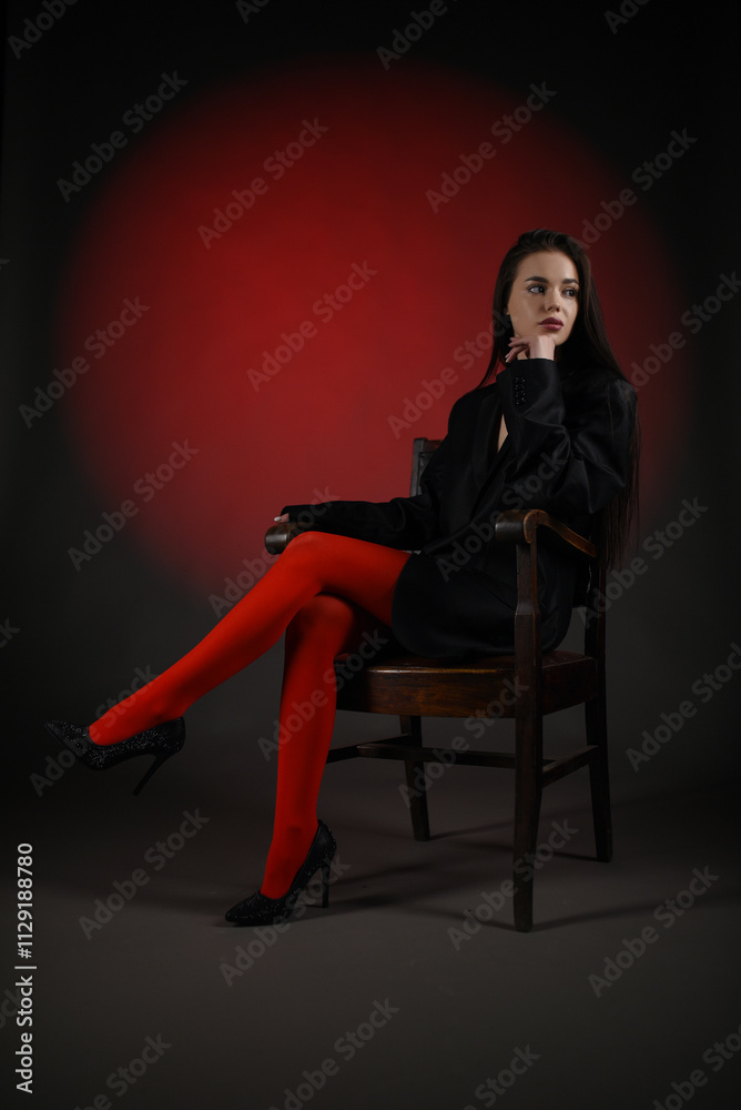 Beautiful girl in a black oversized blazer and red tights posing on the chair on a dark background. Beautiful girl in a black oversized blazer and red tights posing on the chair on a dark background.
