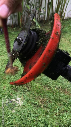 A close-up image of a person cleaning a grass trimmer blade with a brush. The blade is covered in grass clippings, and the image highlights the detail of the cleaning process.