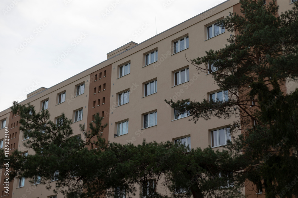 A large apartment building featuring numerous windows and a number of trees situated right in front of it, creating a pleasant view A large apartment building featuring numerous windows and a number of trees situated right in front of it, creating a pleasant view