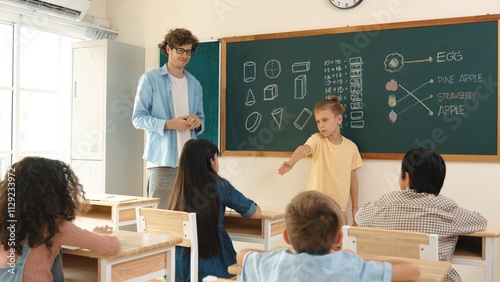 Smart boy pointing at diverse friend while attractive children sitting at class. Elementary student talking and explain idea to caucasian teacher listening while standing in front of class. Pedagogy.