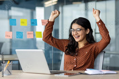 Businesswoman with glasses raising arms in excitement, celebrating achievement at her workplace.