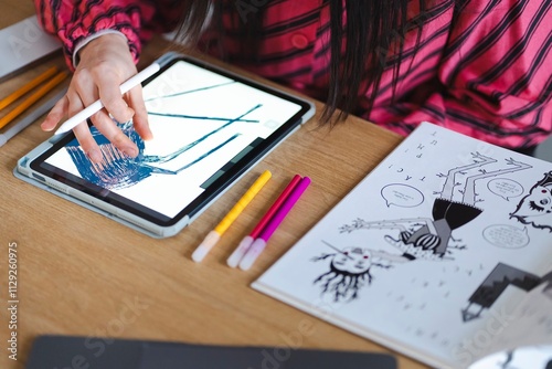 A close-up of a person using a stylus to draw a colorful design on a tablet screen, with colored pens and a comic book on a wooden desk in a creative workspace photo