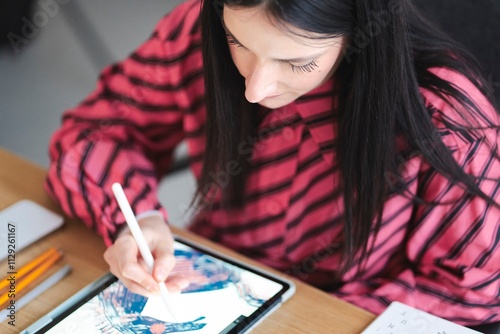 A close-up view of a hand using a stylus to draw intricate patterns on a tablet screen, with colorful pens and a comic book resting on a wooden desk nearby photo