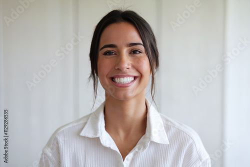 Mujer morena mirando a cámara y sonriendo con una dentadura perfecta. Camisa blanca y fondo blanco. Generado con IA