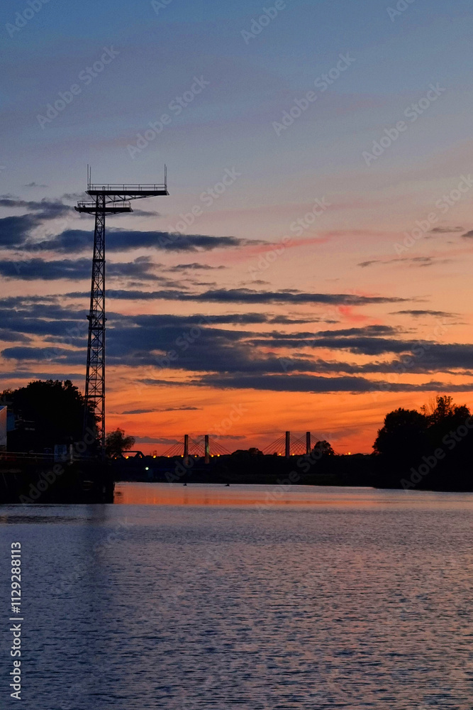 Naklejka premium Serene Sunset over River with Bridge Silhouette and Transmission Tower