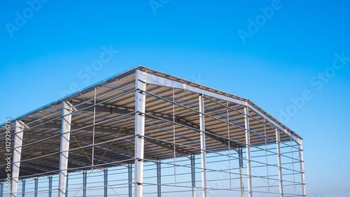 Factory building framework with metal post structure and gable roof in construction site against blue sky background, low angle and perspective side view