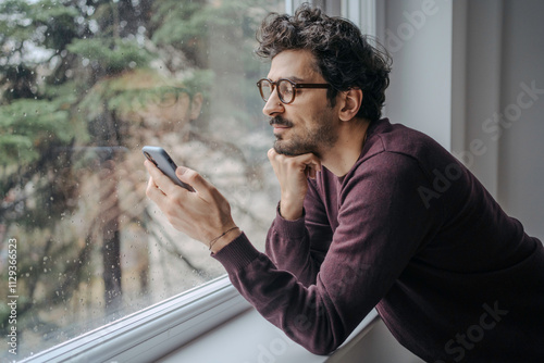 Young handsome middle eastern man scrolling on mobile phone in the morning standing near window.