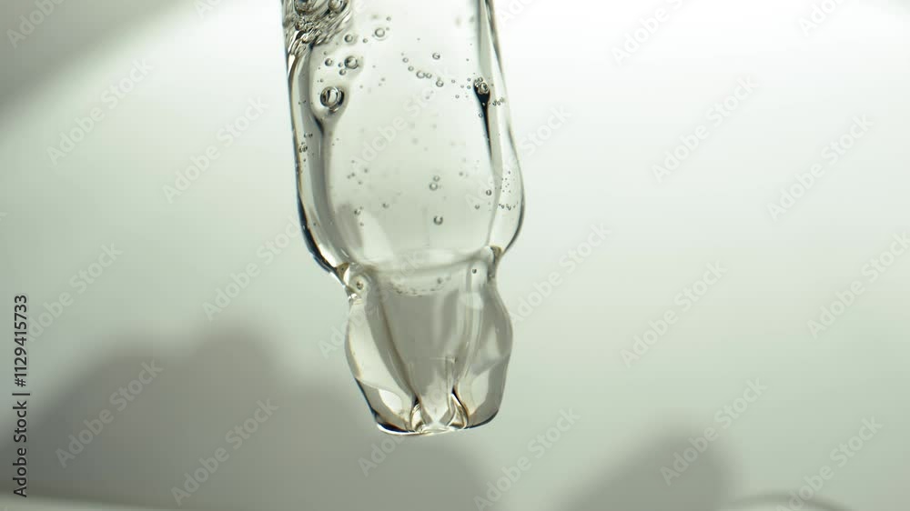 Macro shot of a transparent glass pipette, from which liquid is squeezed out with bubbles in motion against a light background. Concept of cosmetic serum dosage for skin care and beauty maintenance