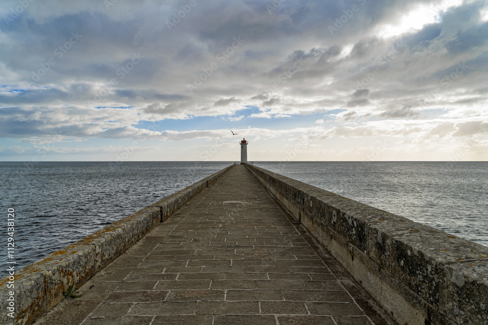 Fototapeta premium Au bout du môle, le phare du Raoulic se dresse sous un ciel nuageux d'automne. Les pavés du môle mènent à lui, créant un contraste saisissant avec le paysage marin environnant.