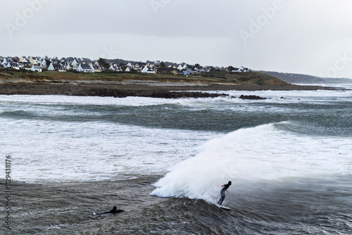 À Audierne, un surfeur brave une vague imposante en pleine tempête. Le ciel nuageux et l’océan déchaîné soulignent son courage face à la puissance des éléments naturels. photo