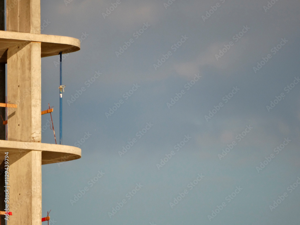 Unfinished building under construction, with a focus on the concrete framework and rebar Part of a crane suggests ongoing work at height Fair weather conditions indicated by a mix of cloudy and blu Unfinished building under construction, with a focus on the concrete framework and rebar Part of a crane suggests ongoing work at height Fair weather conditions indicated by a mix of cloudy and blu