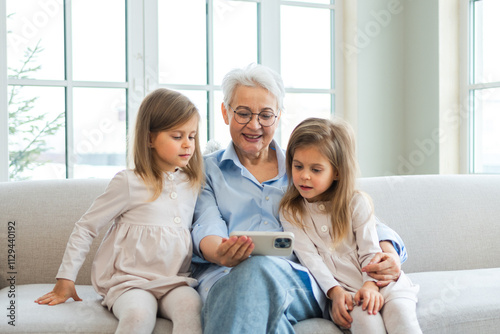 Happy family at home. Two little girls sisters twins grandmother enjoying time together watching video on phone. Good time at home. Grandma granddaughters child kids together. Family generations