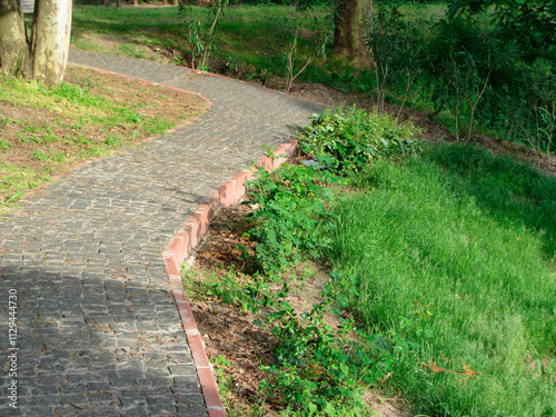 A curved stone pathway with red brick borders, surrounded by lush greenery and grass, set in a park or garden The pathway is aged and structured, photographed during late afternoon or early evening photo