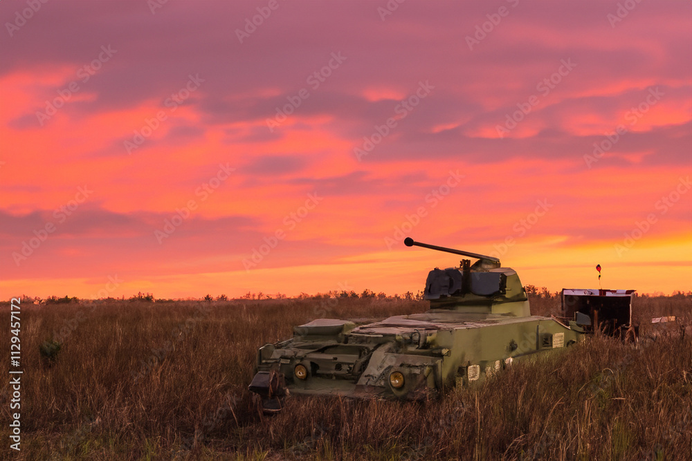 Large tank sits on the grass in front of red sky. A field of tanks at sunset in the evening. A green field with a transport tank at sunset. A green field of tanks at sunset with green lifestyle.