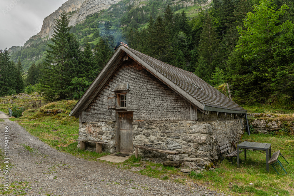 Old stone mountain hut with wooden rooftop in the Appelzeller Alps during a cloudy day in summer. Small Swiss alpine hut, no luxury. Mountain and pine trees in the background. Old stone mountain hut with wooden rooftop in the Appelzeller Alps during a cloudy day in summer. Small Swiss alpine hut, no luxury. Mountain and pine trees in the background.