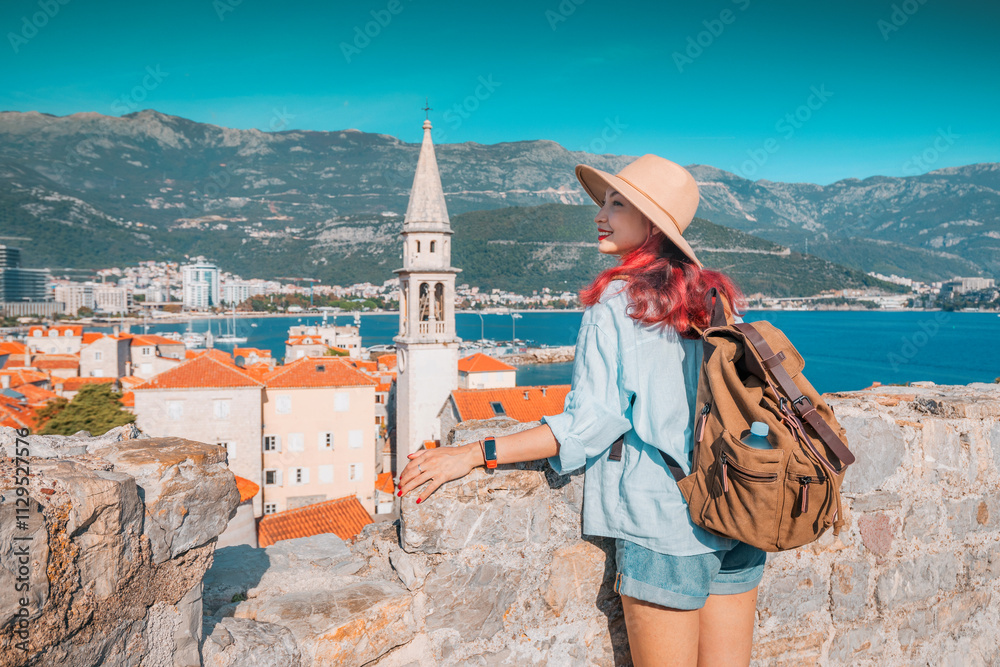 Woman tourist with backpack enjoying Budva old town cityscape in Montenegro from ancient citadel walls with raised arms