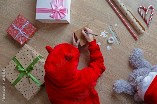 Child Drawing on Gift Box Amid Christmas Prep. Top view of a child in a red hoodie drawing on a small gift box, surrounded by wrapped presents, candy canes, and holiday decorations.