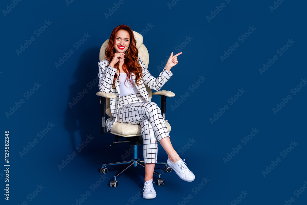 Young woman with red hair in a trendy checkered suit sits on a chair against a dark blue background Young woman with red hair in a trendy checkered suit sits on a chair against a dark blue background