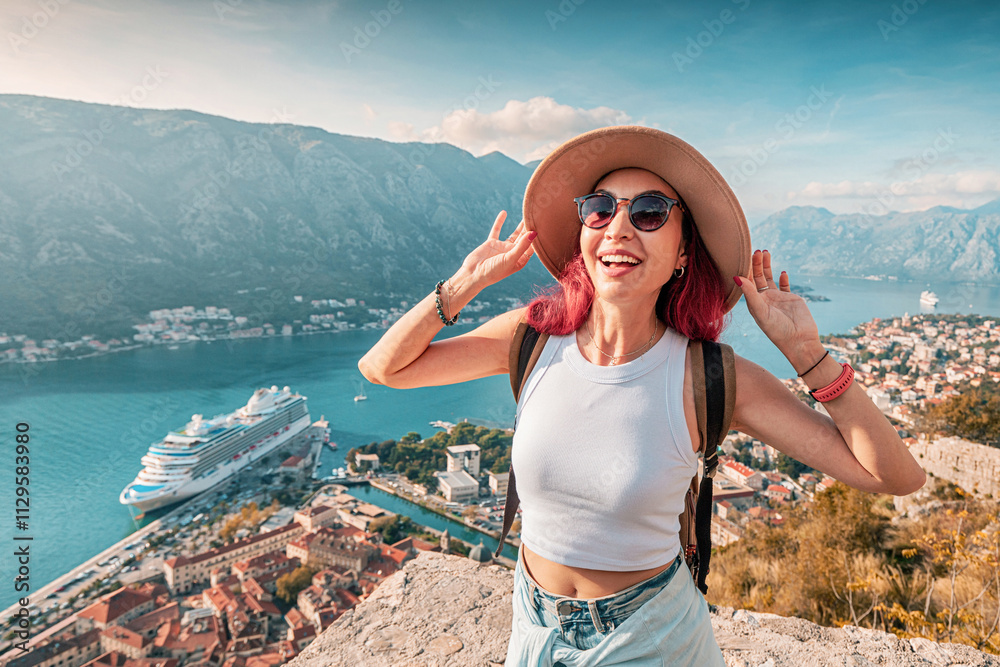 Female tourist with backpack and hat enjoying the view of Kotor bay with cruise ship from fortress