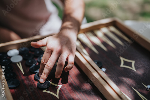 Close-up of a person's hand playing backgammon outdoors, capturing the strategic move in a sunlit setting on a wooden board.