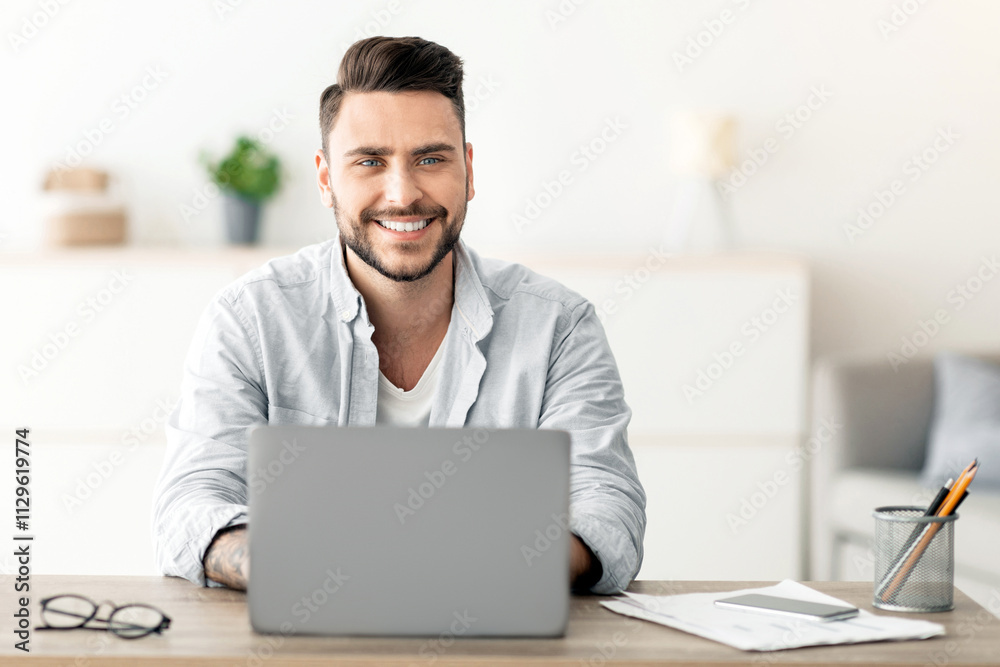 Successful freelancer. Happy man sitting at workplace with laptop computer, smiling and looking at camera, working remotely on computer at home office, copy space. Guy enjoying online distant job Successful freelancer. Happy man sitting at workplace with laptop computer, smiling and looking at camera, working remotely on computer at home office, copy space. Guy enjoying online distant job
