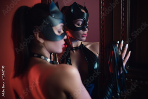 Sexy female lover in latex bdsm costume and leather cat mask posing erotically in front of a mirror in neon light