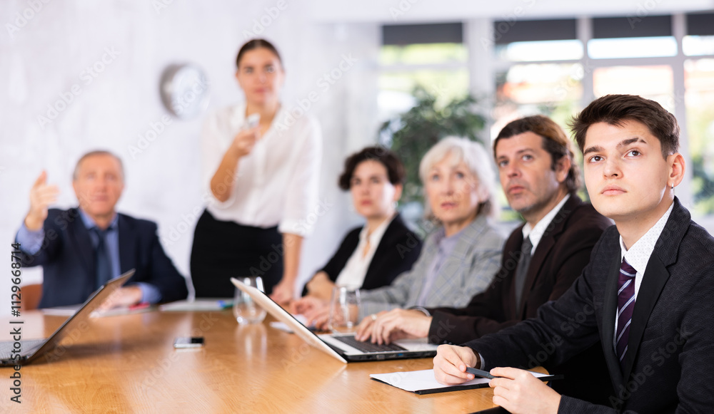 Portrait of young office worker sitting at table with colleagues, listening with interest and watching presentation during business meeting Portrait of young office worker sitting at table with colleagues, listening with interest and watching presentation during business meeting