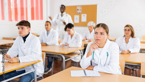 Interested young female student in white coat listening to lecture and taking notes in classroom during professional medical training