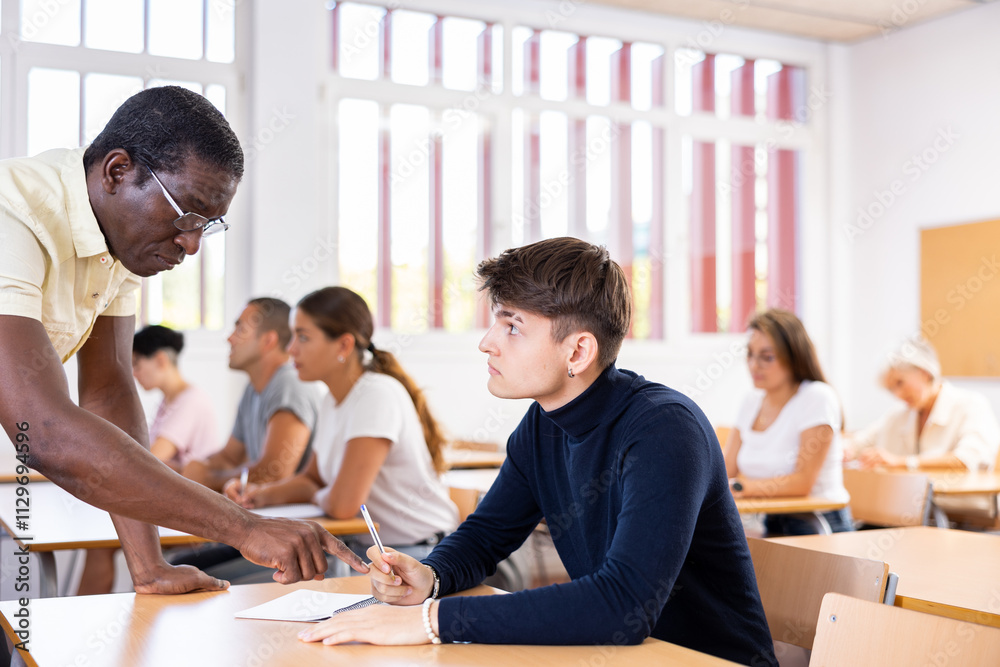 Strict African American teacher standing next to focused young guy sitting at desk during lesson, pointing out error in solving task in student notebook.. Strict African American teacher standing next to focused young guy sitting at desk during lesson, pointing out error in solving task in student notebook..