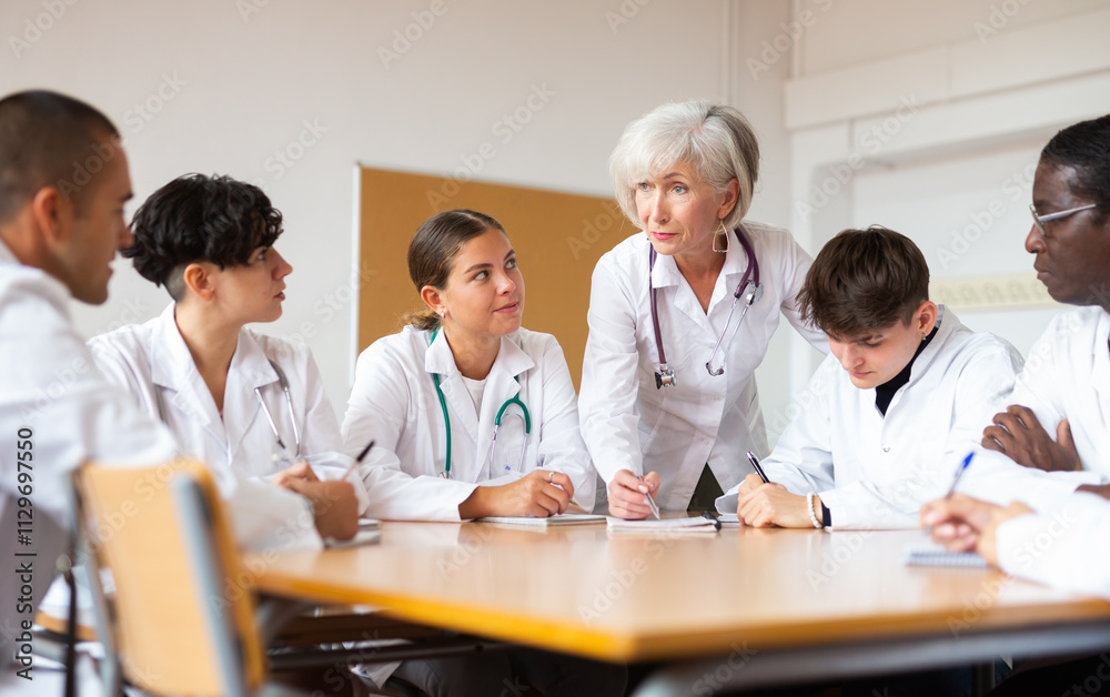 Experienced elderly female professor of medicine giving lecture to group of interested internship doctors sitting at same table in professional seminar.. Experienced elderly female professor of medicine giving lecture to group of interested internship doctors sitting at same table in professional seminar..