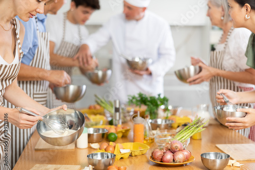 Pleased young woman member of cooking club trying to prepare sauce standing around kitchen table together with others photo