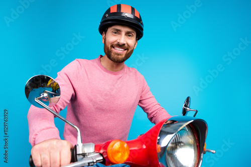 Confident bearded man in a pink sweater smiling while sitting on a red scooter against a bright blue background photo