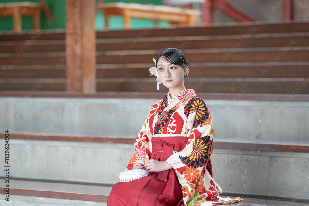 Japanese woman in her 20s wearing red hakama, staple for Japanese university graduates, sits in audience seats in large room. Japanese woman in her 20s wearing red hakama, staple for Japanese university graduates, sits in audience seats in large room.