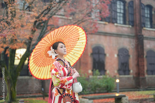 A Japanese woman in her 20s, wearing a red kimono (hakama), a staple of Japanese university graduates, stands in the rain holding a red Japanese umbrella in front of a traditional brick building. photo