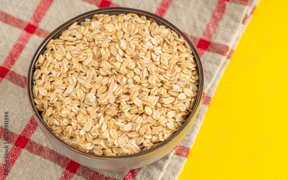 Rustic bowl filled with rolled oats on a checkered cloth with a yellow background. Ideal for breakfast and healthy eating themes. Rustic bowl filled with rolled oats on a checkered cloth with a yellow background. Ideal for breakfast and healthy eating themes.