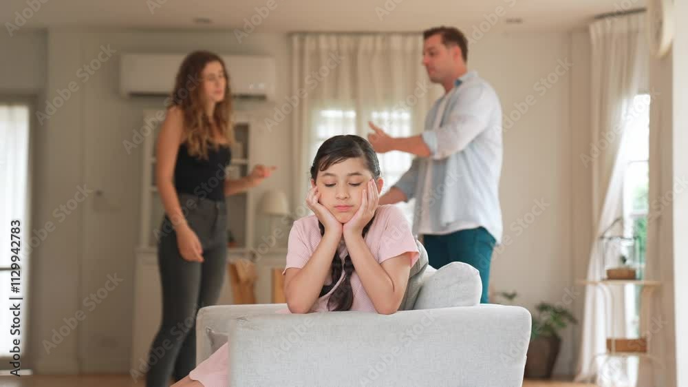 Annoyed and unhappy young girl sitting on sofa trapped in middle of tension by her parent argument in living room. Unhealthy domestic lifestyle and traumatic childhood develop to depression Fastidious Annoyed and unhappy young girl sitting on sofa trapped in middle of tension by her parent argument in living room. Unhealthy domestic lifestyle and traumatic childhood develop to depression Fastidious
