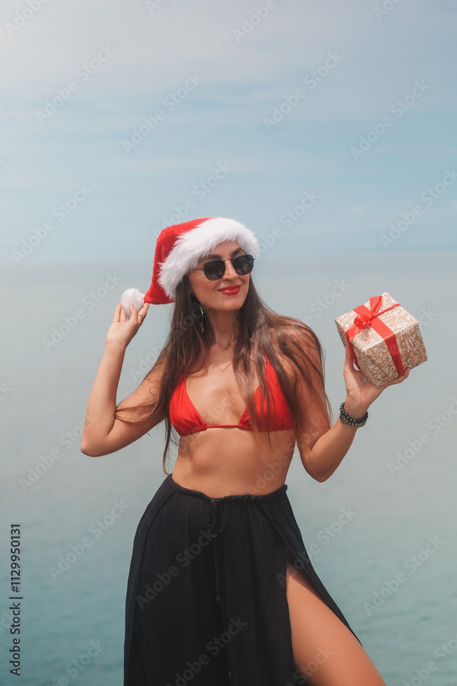 Obraz premium Portrait of young brunette woman in sunglasses, red bikini and Santa hat holding gift box with bow on ocean background. Tropical Christmas and New Year in summer on island. Holiday surprise.