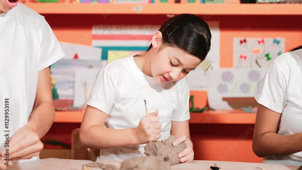 Smart high school girl using carving tool working at clay at pottery workshop. Group of happy diverse children working or modeling cup of clay with dough and equipment scatter around. Edification