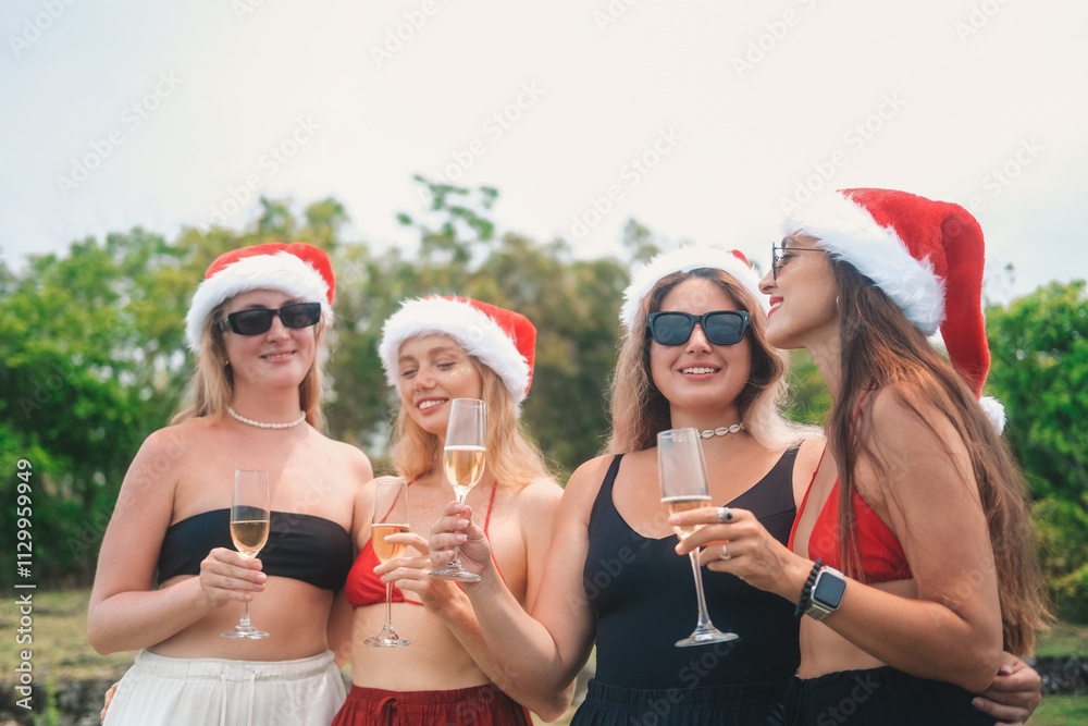 Obraz premium Young women friends stand hugging on a cliff with the ocean view in the background, wearing Santa hats and bikinis. They hold champagne glasses and clink them smiling. Tropical Christmas and New Year.