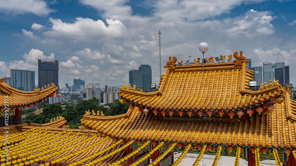 Bright tiled roofs of a Chinese temple against the city skyscrapers, blue sky, clouds. Traditional red and yellow lanterns are suspended along the curved edges. Figures of mythical creatures Bright tiled roofs of a Chinese temple against the city skyscrapers, blue sky, clouds. Traditional red and yellow lanterns are suspended along the curved edges. Figures of mythical creatures