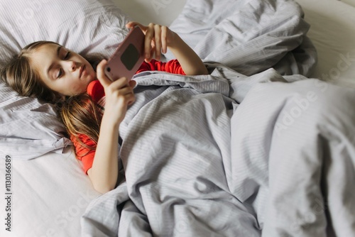 Pretty teenage girl using app on phone, talking on online video call, laughing, secretly watching movie, playing online games, lying under covers on bed. Generation alpha concept. Part of a series