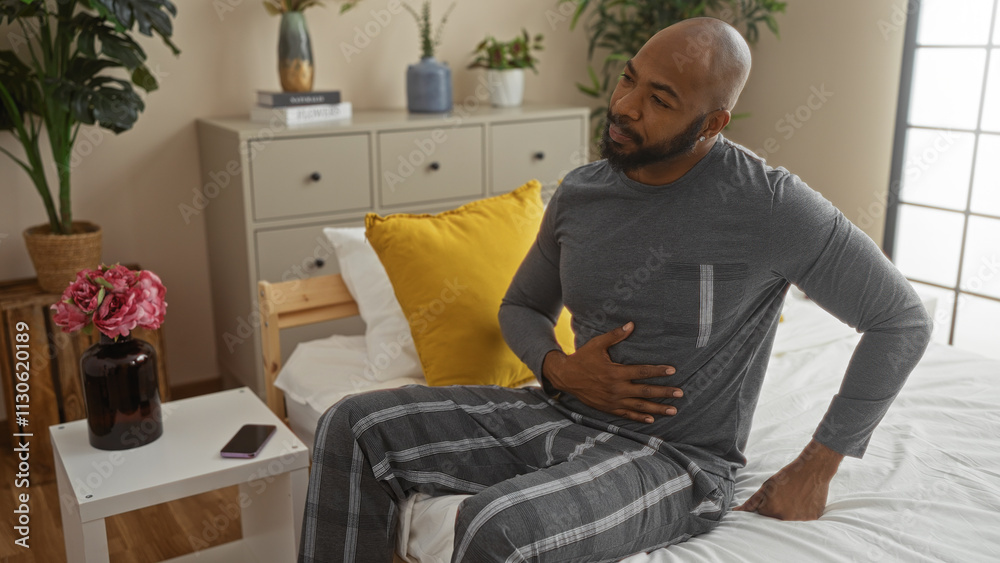 Fototapeta premium Handsome young african american man sitting on bed in cozy bedroom, holding his stomach with a concerned expression, surrounded by indoor plants and a neatly organized room