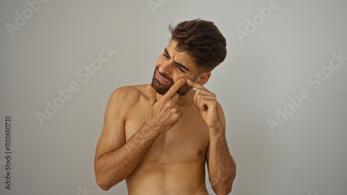Young hispanic man with a beard popping a pimple on his face while standing isolated against a white background.