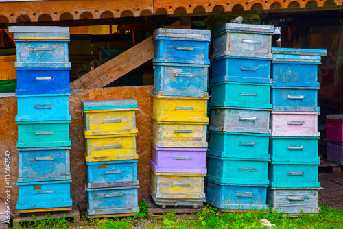 A vibrant array of colorful wooden beehives is stacked under a rustic shelter. The worn paint gives a charming, weathered look, surrounded by greenery, reflecting a serene rural beekeeping scene
