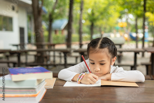 Adorable Asian girl intently writes outdoors, focused on her homework. Surrounded by books, the scene captures childhood learning, concentration, and the joy of studying in outdoor nature.