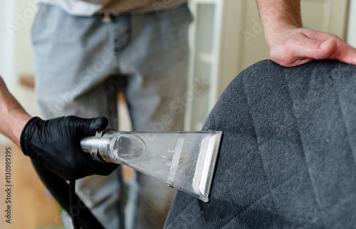 A person wearing gloves is using a vacuum attachment to clean the upholstery of a chair. The bright indoor setting