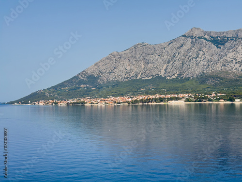 Orebic, Croatia - June 30, 2024: Seen from Adriatic see: The tourist town, red roofed buildings, with its long sandy beach and tall rocky mountain range as backdrop