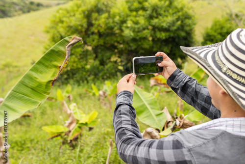 Farmer seen from behind, proud of his land, takes a photograph of his farm with his mobile phone, highlighting a rural landscape with crops and nature